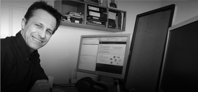 [Photo: Z-omo founder Colin Tester, working at his desk—no fewer than three monitors are required.]